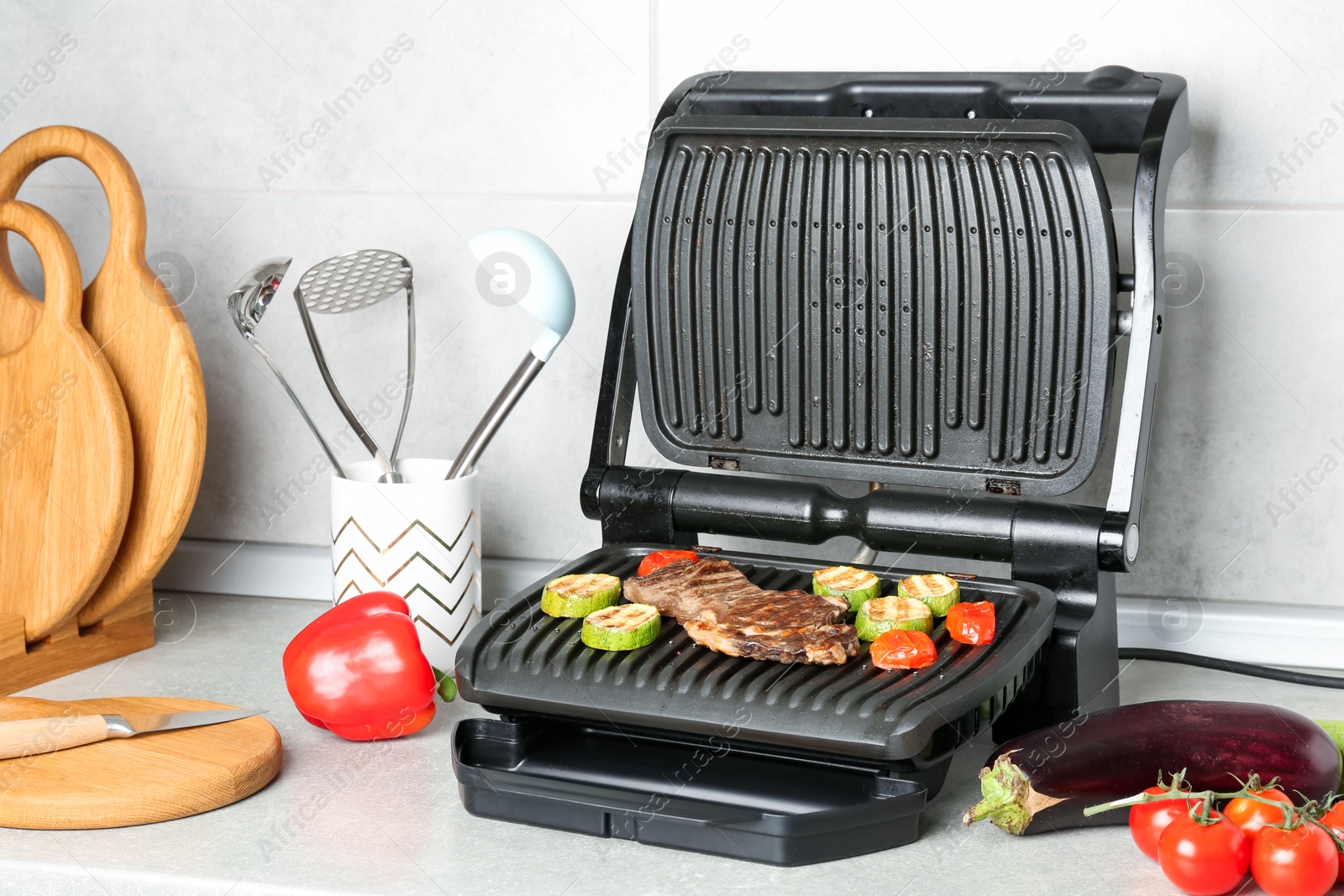 Photo of Open electric grill with beef steak, vegetables and cookware on light grey table