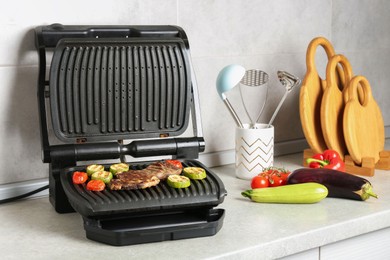 Photo of Open electric grill with beef steak, vegetables and cookware on light grey table