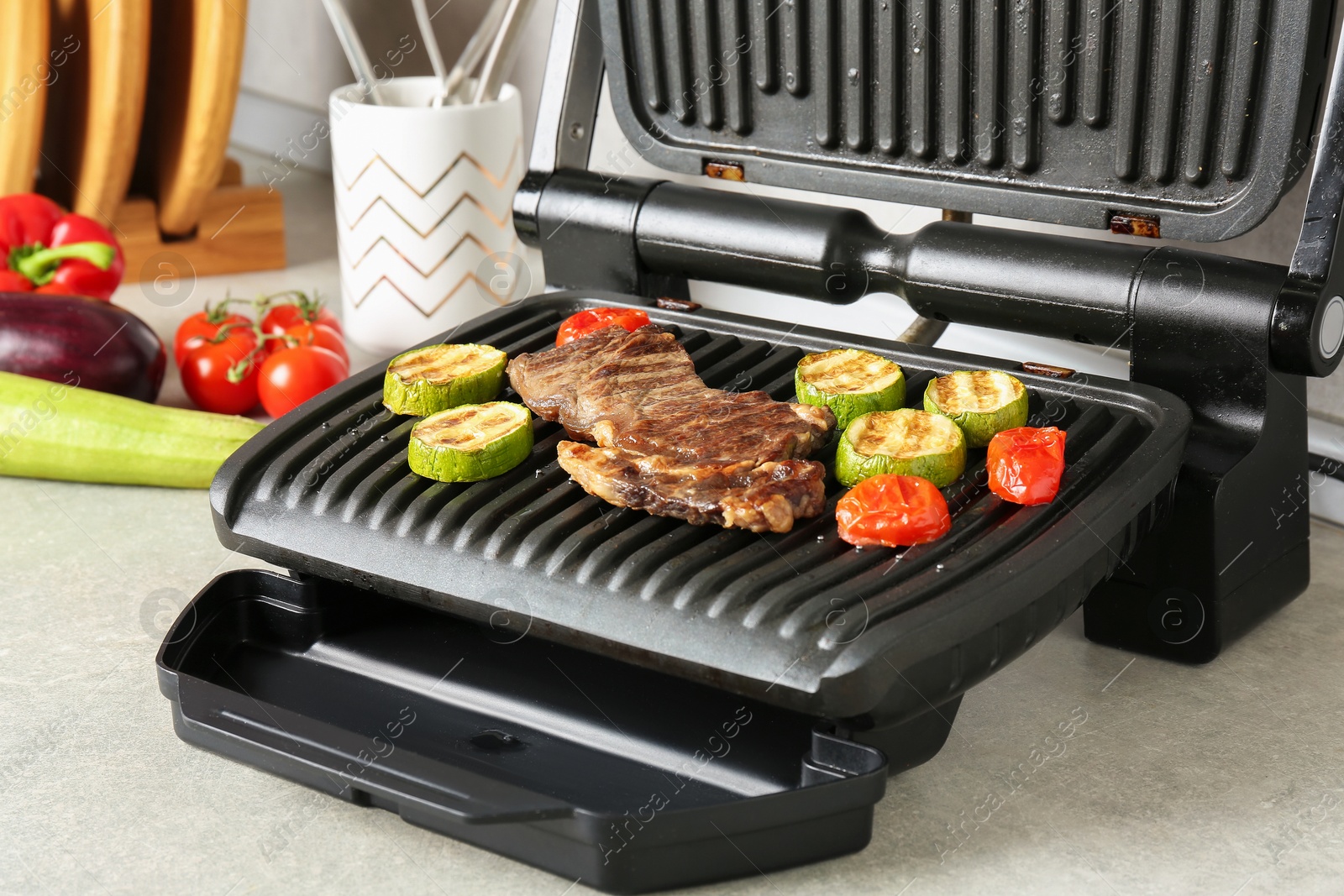 Photo of Open electric grill with beef steak, zucchini and tomatoes on light grey table, closeup