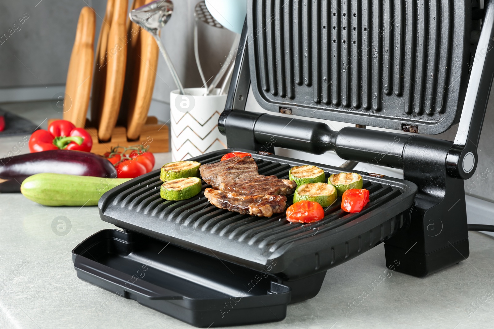 Photo of Open electric grill with beef steak and vegetables on light grey table, closeup