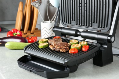 Photo of Open electric grill with beef steak and vegetables on light grey table, closeup