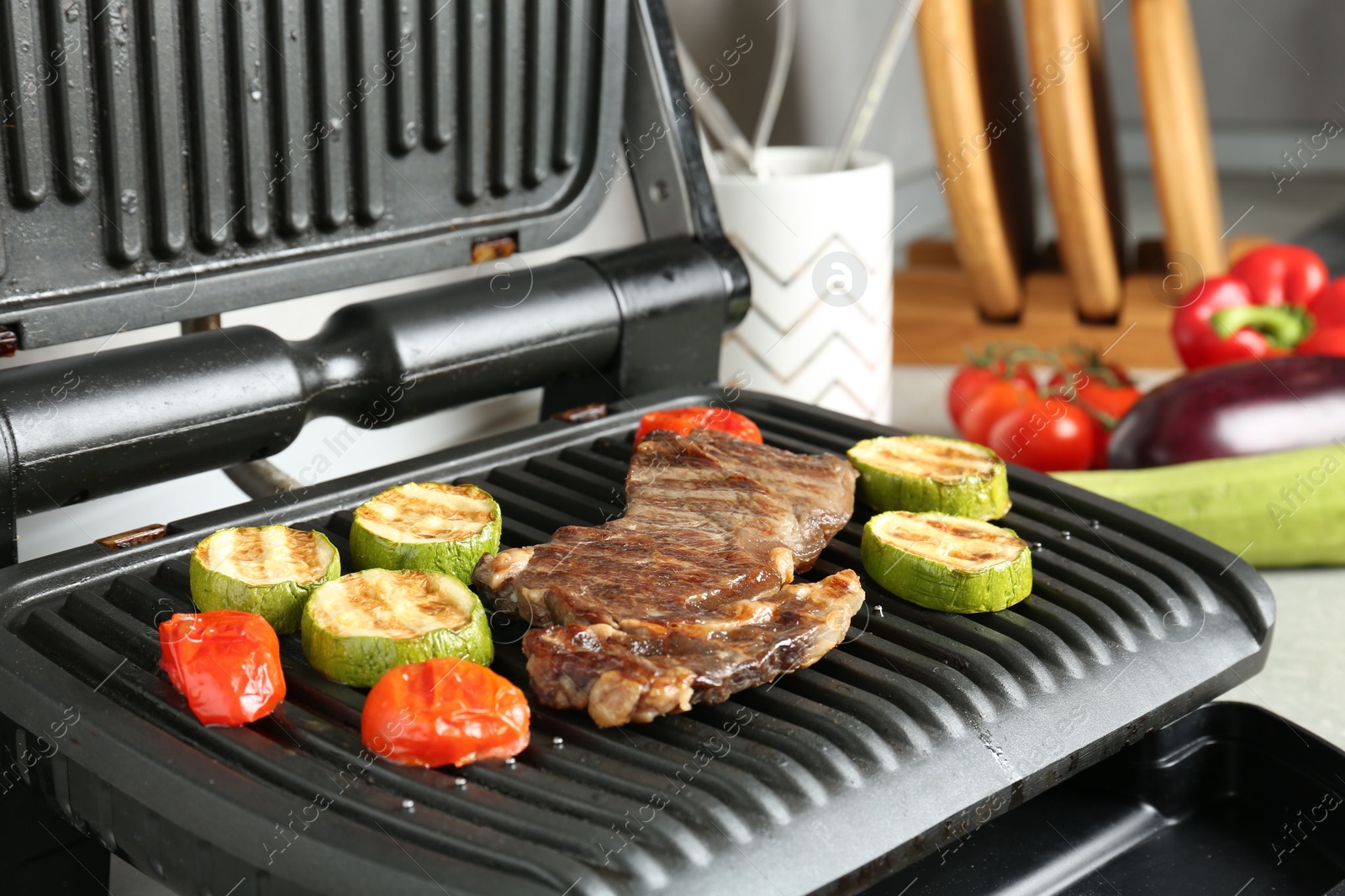 Open electric grill with beef steak, zucchini and tomatoes on table, closeup Photo of Open electric grill with beef steak, zucchini and tomatoes on table, closeup