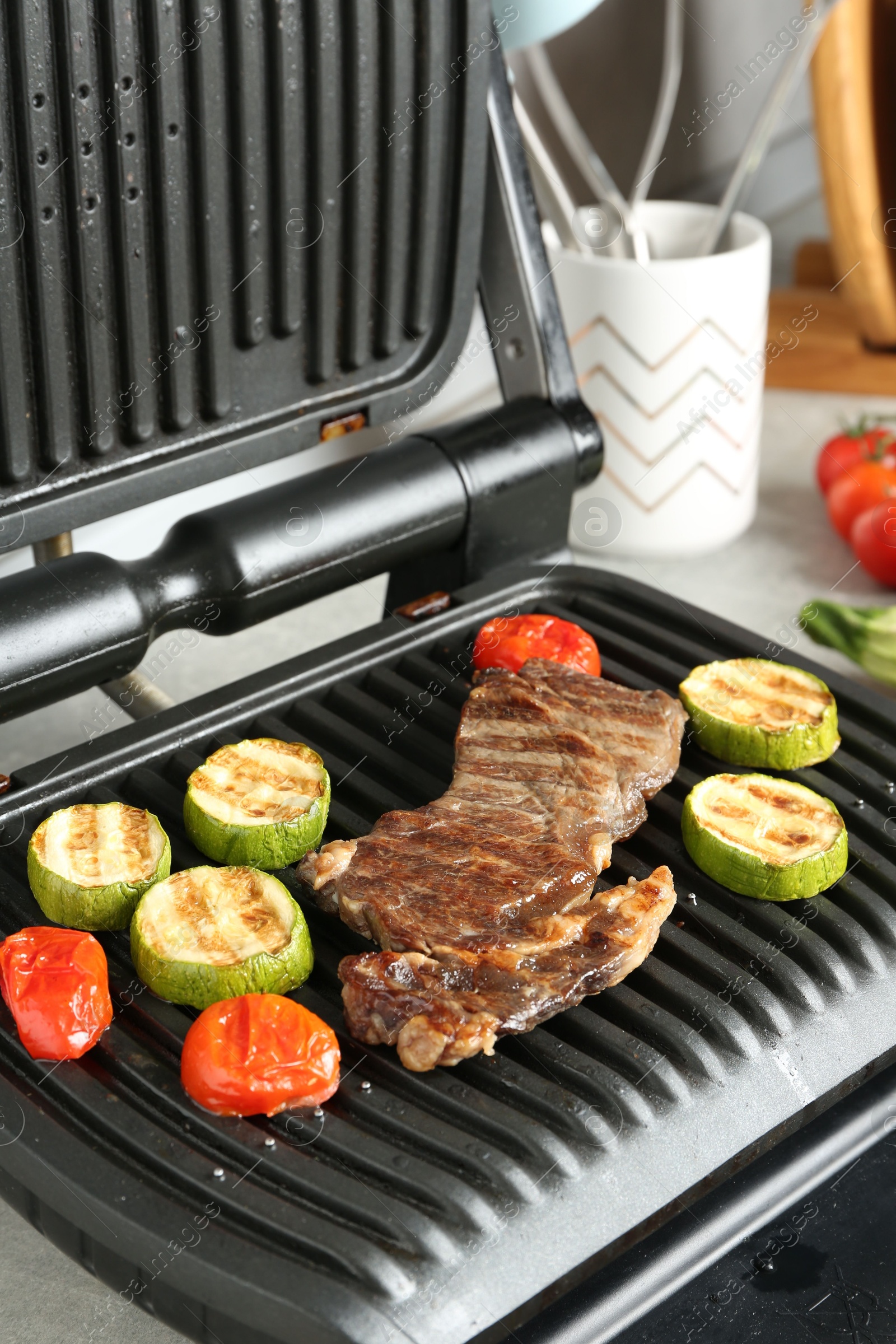 Open electric grill with beef steak, zucchini and tomatoes on table, closeup Photo of Open electric grill with beef steak, zucchini and tomatoes on table, closeup