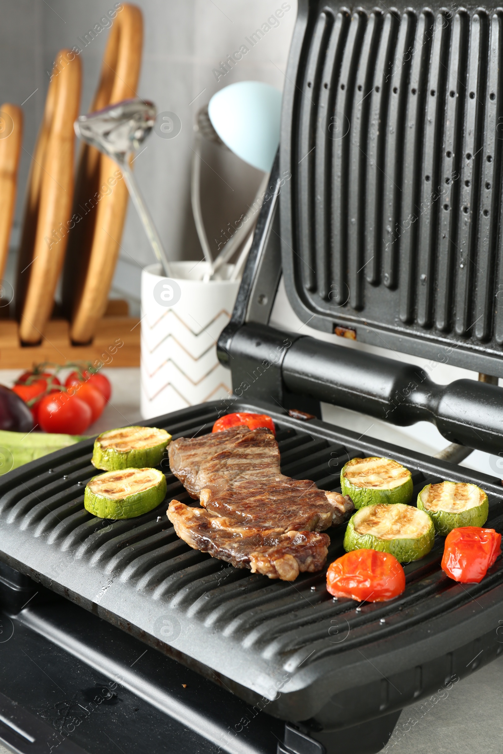 Open electric grill with beef steak, zucchini and tomatoes on light grey table, closeup Photo of Open electric grill with beef steak, zucchini and tomatoes on light grey table, closeup