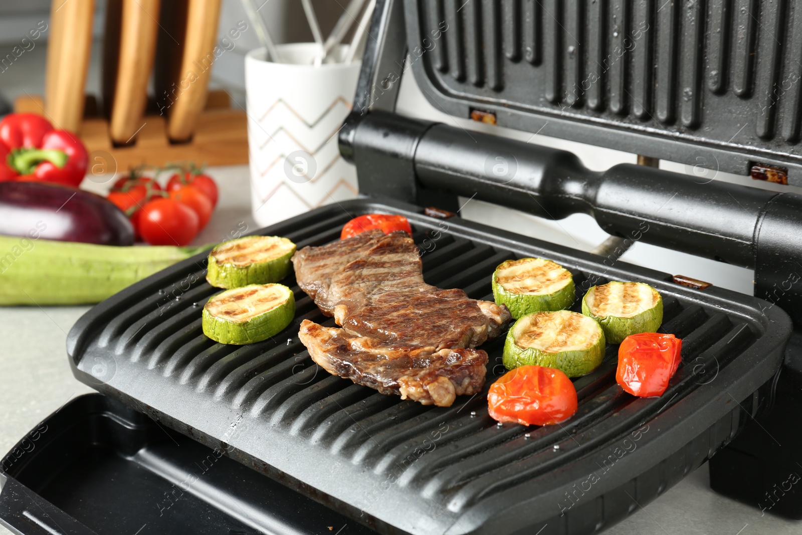 Open electric grill with beef steak, zucchini and tomatoes on light grey table, closeup Photo of Open electric grill with beef steak, zucchini and tomatoes on light grey table, closeup