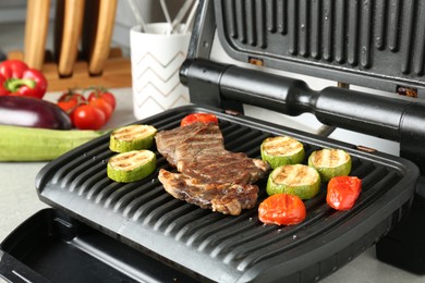 Photo of Open electric grill with beef steak, zucchini and tomatoes on light grey table, closeup