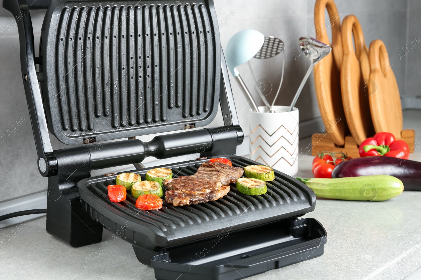 Photo of Open electric grill with beef steak, vegetables and cookware on light grey table, closeup