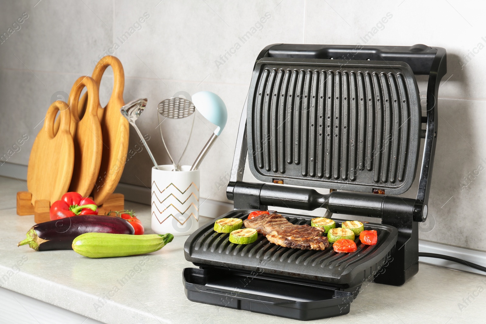 Photo of Open electric grill with beef steak, vegetables and cookware on light grey table