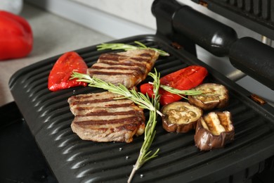 Photo of Open electric grill with beef steaks, rosemary and vegetables on table, closeup