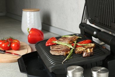 Photo of Open electric grill with beef steaks, rosemary and vegetables on light grey table, closeup