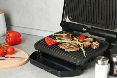 Photo of Open electric grill with beef steaks, rosemary, vegetables and spices on light grey table, closeup