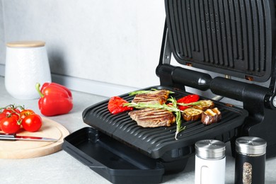 Photo of Open electric grill with beef steaks, rosemary, vegetables and spices on light grey table, closeup