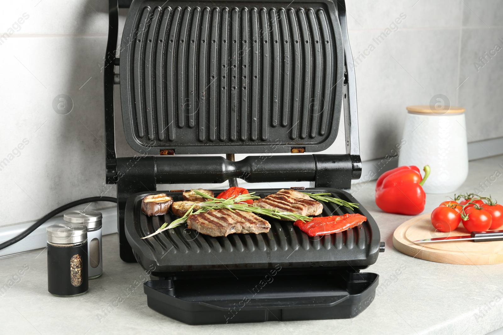 Open electric grill with beef steaks, rosemary, vegetables and spices on light grey table, closeup Photo of Open electric grill with beef steaks, rosemary, vegetables and spices on light grey table, closeup