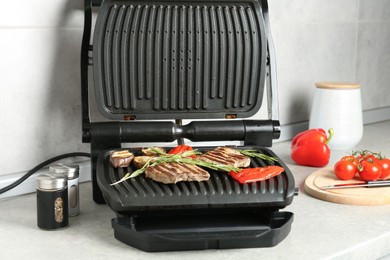 Photo of Open electric grill with beef steaks, rosemary, vegetables and spices on light grey table, closeup