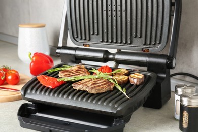 Photo of Open electric grill with beef steaks, rosemary, vegetables and spices on light grey table, closeup