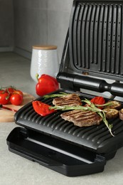 Photo of Open electric grill with beef steaks, rosemary, vegetables and spices on light grey table, closeup