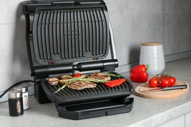 Photo of Open electric grill with beef steaks, rosemary, vegetables and spices on light grey table, closeup