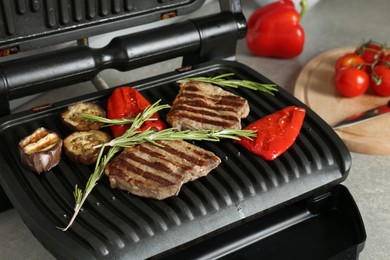 Photo of Open electric grill with beef steaks, rosemary, bell pepper and eggplant on light grey table, closeup