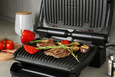 Photo of Open electric grill with beef steaks, rosemary, bell pepper and eggplant on light grey table, closeup
