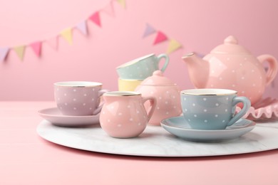 Photo of Beautiful ceramic tea set for children on wooden table against pink background with party flags, closeup