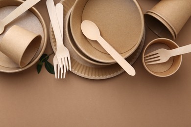 Photo of Set of disposable tableware and green leaves on brown background, flat lay. Space for text