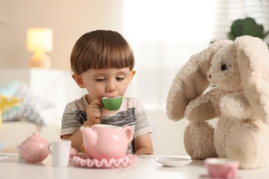 Photo of Cute little boy playing tea party with his toy bunny at table indoors