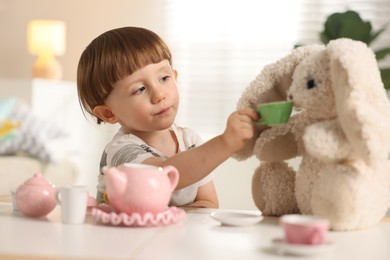Photo of Cute little boy playing tea party with his toy bunny at table indoors
