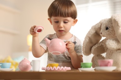 Photo of Cute little boy playing tea party with his toy bunny at table indoors