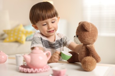 Photo of Cute little boy playing tea party with his toy bear at table indoors