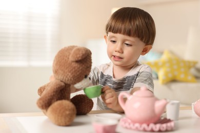 Photo of Cute little boy playing tea party with his toy bear at table indoors