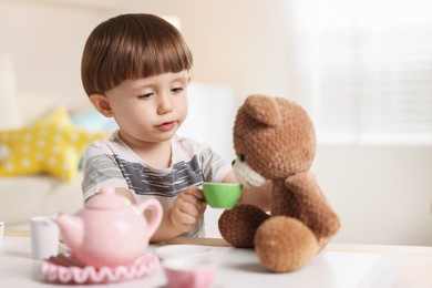 Photo of Cute little boy playing tea party with his toy bear at table indoors