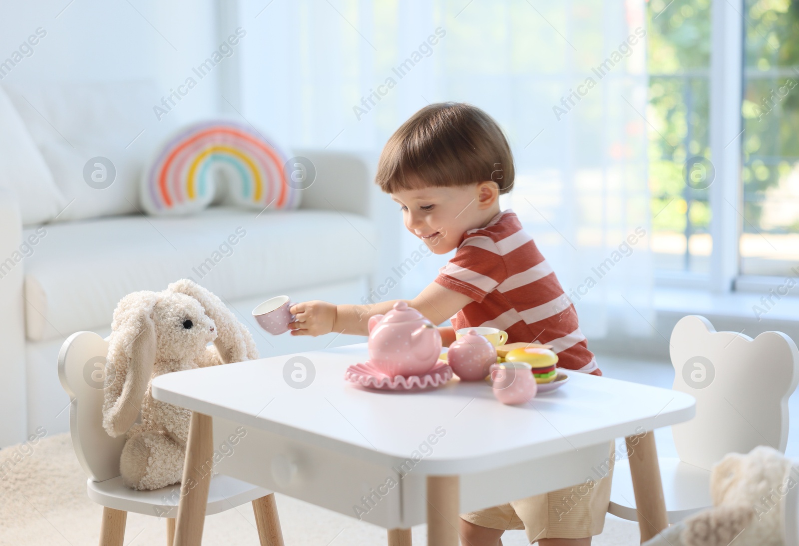 Photo of Cute little boy playing tea party with his toy bunny at table indoors