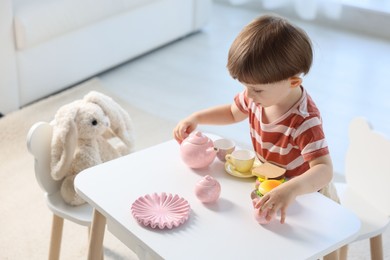 Photo of Cute little boy playing tea party with his toy bunny at table indoors