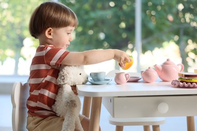 Photo of Cute little boy playing tea party with his toy sheep at table indoors