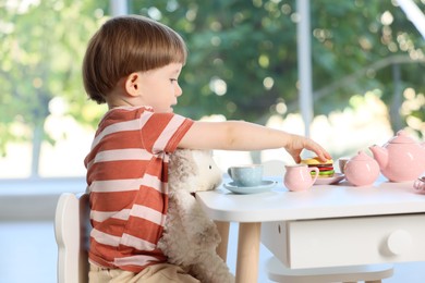 Photo of Cute little boy playing tea party with his toy sheep at table indoors