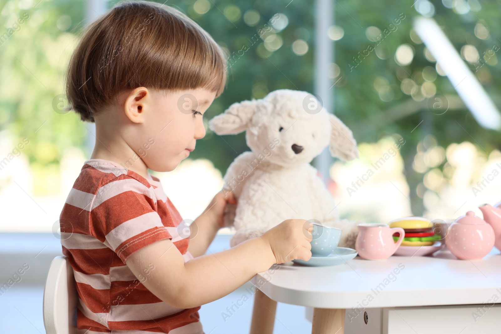 Photo of Cute little boy playing tea party with his toy sheep at table indoors