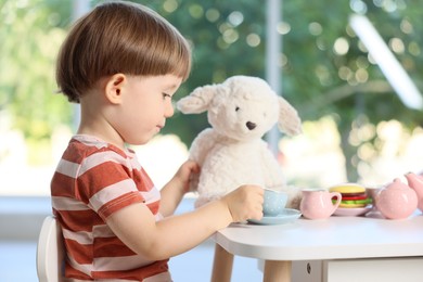 Photo of Cute little boy playing tea party with his toy sheep at table indoors
