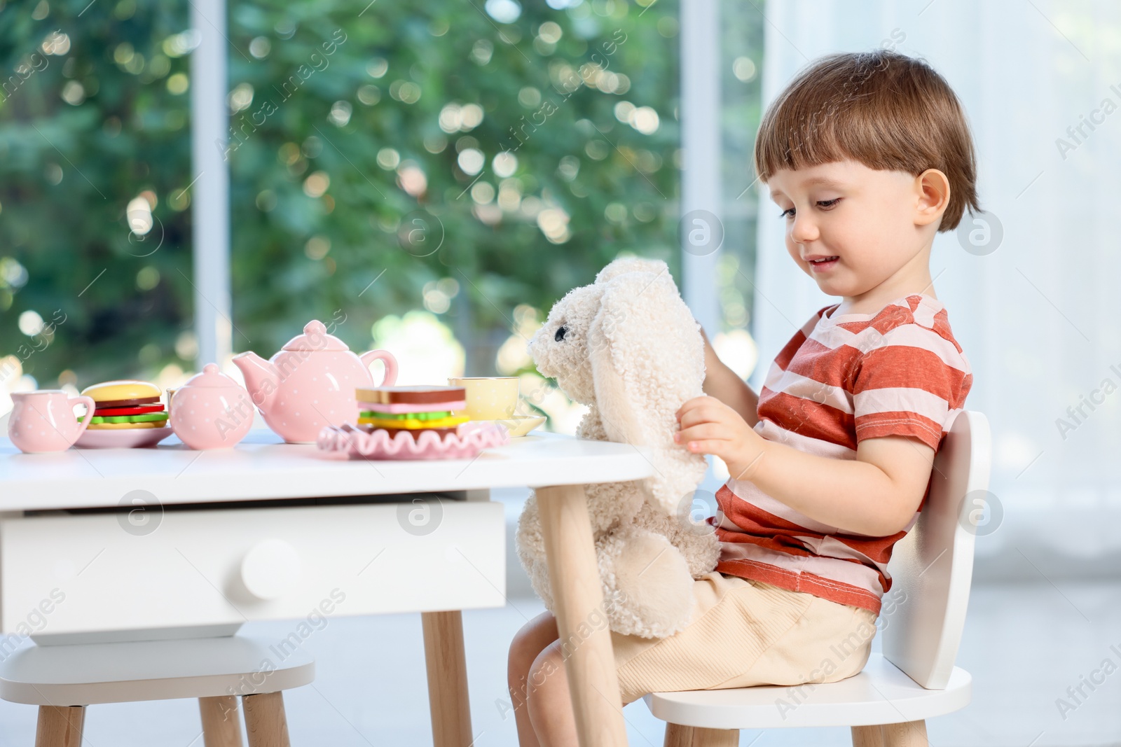Photo of Cute little boy playing tea party with his toy bunny at table indoors