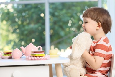 Photo of Cute little boy playing tea party with his toy bunny at table indoors. Space for text