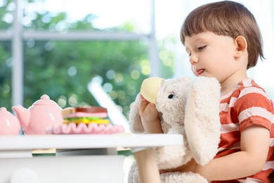 Photo of Cute little boy playing tea party with his toy bunny at table indoors. Space for text