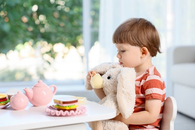 Photo of Cute little boy playing tea party with his toy bunny at table indoors. Space for text