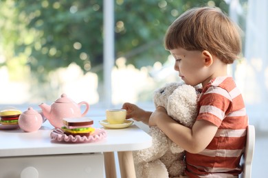 Photo of Cute little boy playing tea party with his toy bunny at table indoors. Space for text