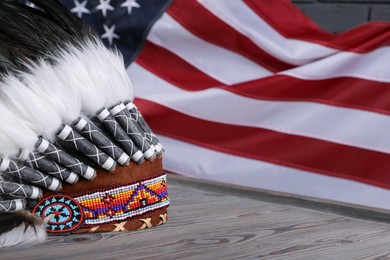 Photo of Native American chief headdress (feathered war bonnet) on wooden surface against American flag, closeup. Space for text