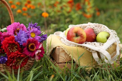 Photo of Beautiful bouquet of flowers in wicker basket and apples on green grass outdoors, closeup