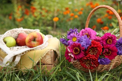 Photo of Beautiful bouquet of flowers in wicker basket and apples on green grass outdoors, closeup