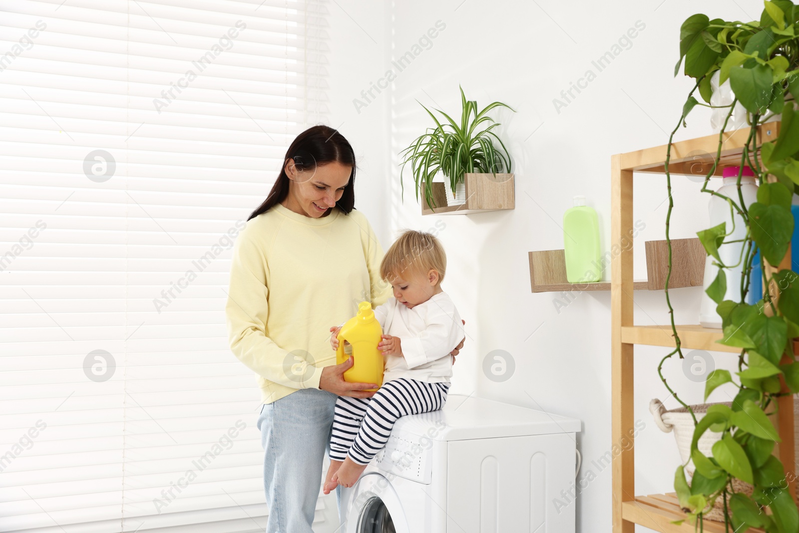 Photo of Smiling housewife and her baby with detergent on washing machine near white wall at home