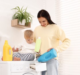 Photo of Smiling housewife and her baby with detergents doing laundry on washing machine near white wall at home