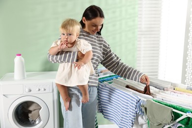 Photo of Smiling mother with her little daughter hanging clean laundry on drying rack at home