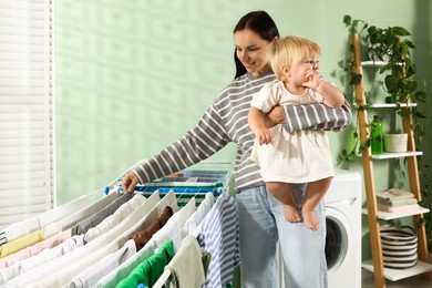 Photo of Smiling mother with her little daughter hanging clean laundry on drying rack at home