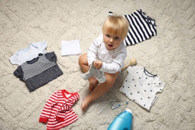 Photo of Cute baby among clean clothes and detergent on light carpet, above view